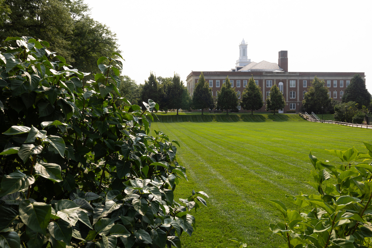 The UNO Pep Bowl, a large green lawn, is pictured in the foreground with the Arts and Sciences Hall in the background, partially framed by leafy trees and bushes. 